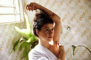 mulher branca de cabelos pretos dentro de um banheiro de arquitetura dos anos 80 e uma samabaia atrás, enrolada em uma toalha com a mão direita na axila esquerda